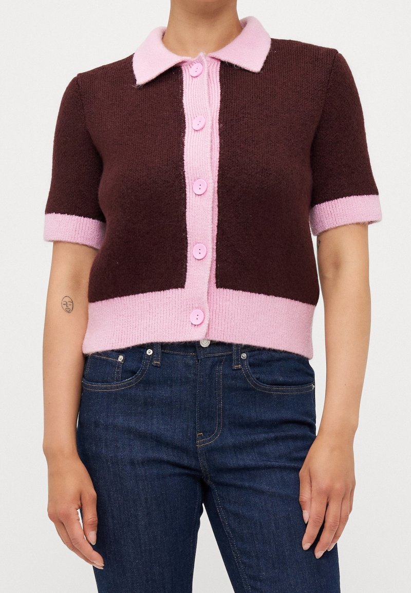 Cardigan marron à manches courtes avec col rose, patte de boutonnage, poignets et ourlet, porté avec un jean bleu foncé.