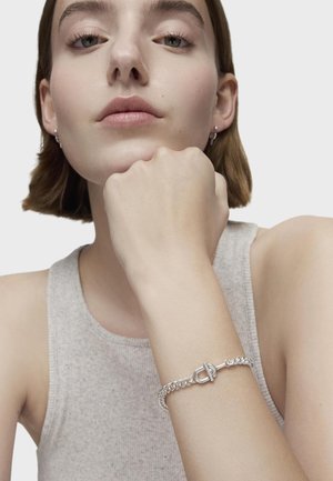 Bracciale in argento con chiusura a toggles, indossato su un polso. La modella ha i capelli corti e indossa una canotta grigia senza maniche. La pelle appare liscia.