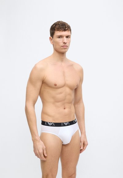 Särgivaba mees, kellel on lühikesed lokkis juuksed, valge musta vöökohaga aluspüksid, millel on EA logo, seisab ühevärvilise hele tausta ees.