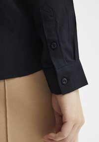 Camicia nera con bottoni, a texture liscia, dotata di due bottoni sui polsini e di un design delle maniche su misura, su uno sfondo beige.