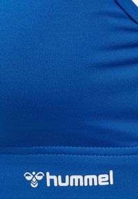 Blå sportbh gjord av ribbat tyg, med en mjuk textur och vit logga nederst. Utformad för komfort och passform.