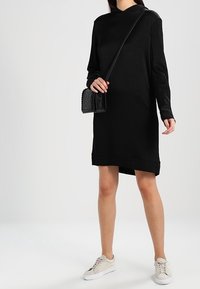Abito nero a maniche lunghe con una vestibilità morbida e colletto alto, abbinato a una borsa a tracolla nera dalla texture interessante e sneakers beige. Design semplice.