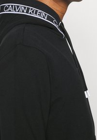 Felpa con cappuccio in cotone nero con cordino, caratterizzata da una fascia con logo bianco lungo il colletto e punte a contrasto sui cordini.