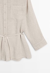 Beige textured long-sleeve button-up shirt with chest pocket and adjustable fabric belt tied at the waist on a white background.