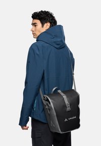 Zwart-grijze Vaude boodschappentas met een gestructureerd oppervlak, verstelbare schouderband en een sluiting aan de voorkant met gesp; gedragen over een blauwe jas.