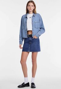 Veste en jean bleu clair, avec deux poches avant, portée sur un haut court blanc. Associée à une jupe en jean bleu foncé taille haute.