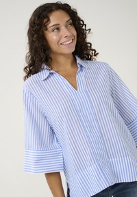 Mėlynos ir baltos juostelės marškiniai su V formos iškirpte, trumpomis rankovėmis ir laisvu kirpimu. Medvilnės audinys su švelniu tekstūra ir šoninėmis įpjovomis patogumui.