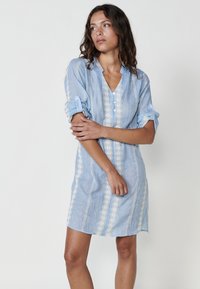 Hellblaues Leinenkleid mit V-Ausschnitt, Knopfblende und vertikalen Spitzenbordüren entlang der Vorderseite und Ärmel.