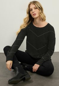 Maglietta nera a maniche lunghe con scollatura rotonda, caratterizzata da un motivo a zig-zag di borchie bianche. Indossata con jeans neri e stivaletti.