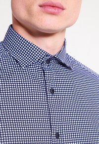 Mörkblå rutiga skjorta med spridd krage, med ett geometriskt mönster i vitt och ljusblått, samt svarta knappar längs knäppningen.