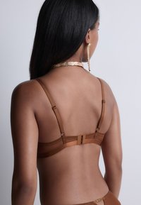 Soutien-gorge marron avec des bretelles ajustables, fermeture à crochets et œillets, et texture lisse ; présente un accent décoratif en métal doré.