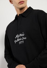 Sort poloshirt med klassisk krave, lange ærmer og hvidt broderet tekst med "Marais Gallerie Deux 1973" detaljer.