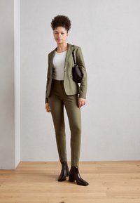 Olivově zelený sak a slim kalhoty v odpovídající barvě, doplněné o bílé tričko. Černá kabelka a kotníkové boty dokončují outfit. Pozadí je dřevěná podlaha.