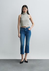 Noor naine seisab seina ääres, kandes triibulist varrukateta kõrge kaelusega pluusi, lühendatud siniseid teksapükse ja musta terava ninaga kontsakingi.