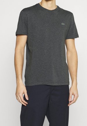 Mand iført en ensfarvet mørkegrå kortærmet t-shirt med lille grønt logo på brystet og mørke bukser, der står mod en hvid baggrund.