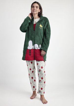Mujer con una chaqueta verde de felpa con cremallera, con diseños de Snoopy y Woodstock, y pantalones de pijama blancos con temática de Peanuts, de pie descalza.
