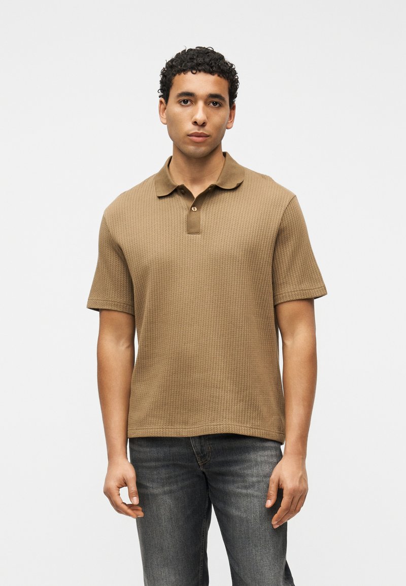 Homme aux cheveux noirs bouclés portant un polo marron à manches courtes texturé et un jean foncé délavé, debout devant un fond blanc uni.