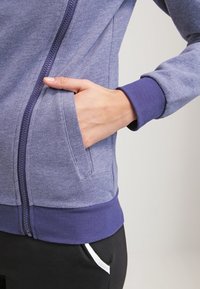 Giacca a zip viola con una finitura testurizzata, dotata di una tasca frontale e polsini a costine. Completata da una vita solida in una tonalità più scura.
