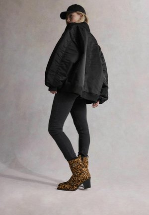 Jachetă bomber neagră peste blugi skinny negri. Botine cu imprimeu leopard tare, maro, și tocuri groase. Model purtând o șapcă neagră.
