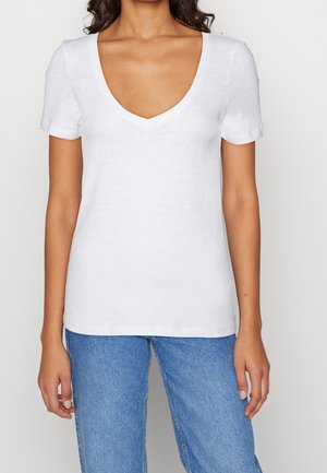 Kvinde iført en simpel hvid T-shirt med korte ærmer og V-hals samt blå jeans, vist fra skuldre til hofter mod en ensfarvet baggrund.