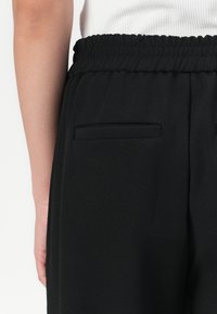 Zwarte broek met een zachte, gladde textuur. Heeft een elastische tailleband en twee decoratieve achterzakken.
