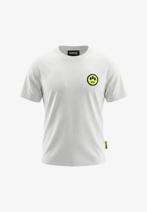 T-shirt bianca a maniche corte con scollo rotondo, caratterizzata da un piccolo smiley giallo-verde con tre occhi e zanne sul lato sinistro del petto.