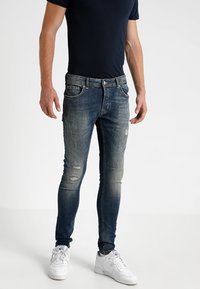 Man med åtsittande mörkblå slitna jeans, svart kortärmad skjorta och vita sneakers, stående mot en neutral bakgrund.