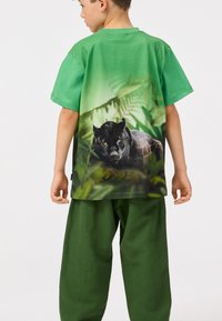 Zeleni pamučni majica s velikim crnim grafičkim prikazom pantere na leđima, u kombinaciji s labavim zelenim hlačama.