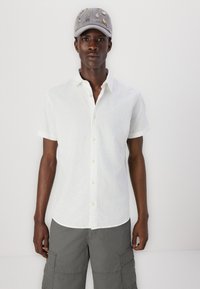 Weißes kurzärmliges Leinenhemd mit Button-Down-Kragen, kombiniert mit grauen Cargo-Shorts. Graue Kappe, verziert mit verschiedenen bunten Aufnähern.