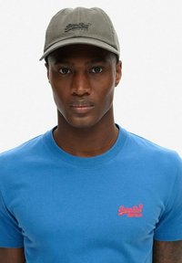 Grijze honkbalpet met geborduurd logo, gecombineerd met een blauwe ronde hals t-shirt met een klein roze logo op de borst.