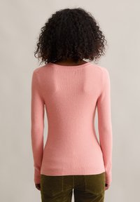 Frau mit lockigem Haar trägt ein eng anliegendes, rosa, geripptes Langarmshirt und olivgrüne Cordhosen, steht mit dem Rücken zur Kamera vor einem neutralen Hintergrund.