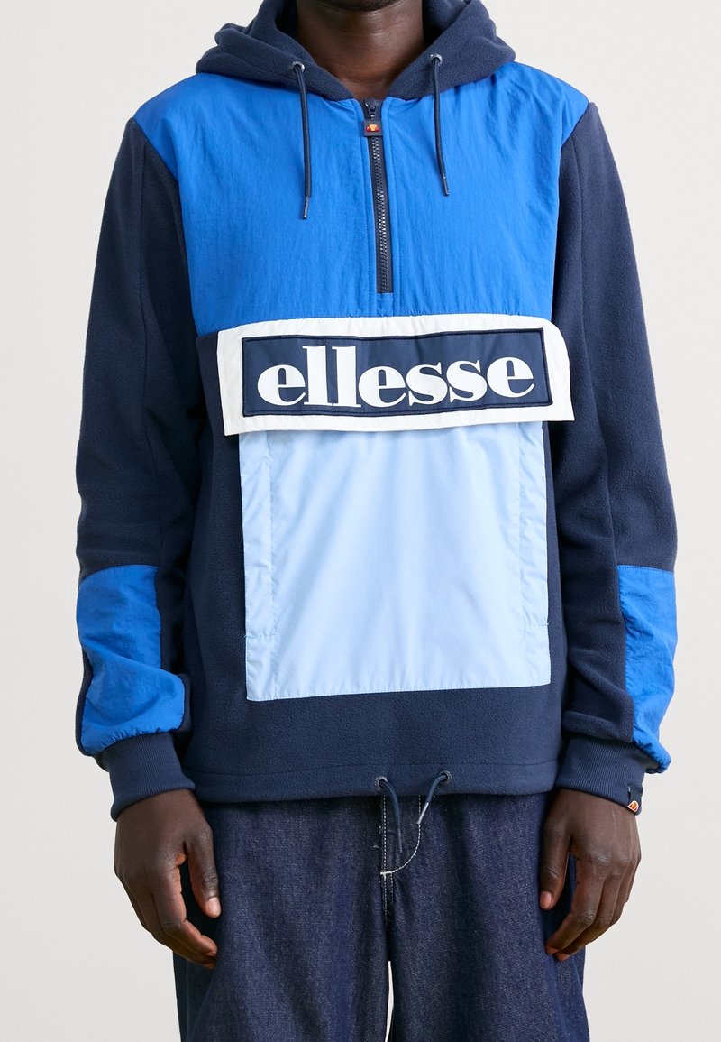 Blå och marinblå huvtröja med halv dragkedja, utrustad med en ljusblå panel och en framträdande vit logotyp över bröstet. Mjuk textur.