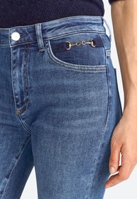 Jean en denim bleu foncé avec une coupe slim, accents en métal doré, et une taille texturée avec un détail de chaîne décorative.