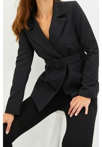 Musta bleiseri, jossa vyötäröllä vyö ja pyöreä solki, shaalikaulus ja ryhdikkäät olkapäät. Sileä kangas, räätälöity istuvuus.