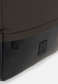 Donkerbruine stoffen tas met een zwarte lederen basis, met een rechthoekige vorm en geperst logo-detail, gladde textuur en minimalistisch ontwerp.