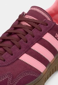 Μπορντό σουέτ αθλητικό παπούτσι με ροζ λεπτομέρειες και τρεις ρίγες. Διαθέτει υφασμένο πέλμα και καφέ κορδόνια. Ραφές κατά μήκος των άκρων.