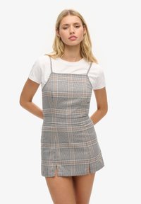 Junge Frau, die ein graues karierte ärmelloses Minikleid über einem weißen Kurzarm-T-Shirt trägt und mit Händen hinter ihrem Rücken steht.