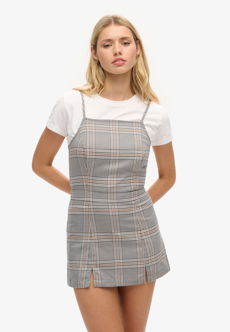 Junge Frau, die ein graues karierte ärmelloses Minikleid über einem weißen Kurzarm-T-Shirt trägt und mit Händen hinter ihrem Rücken steht.