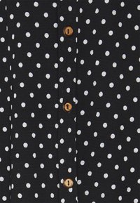 Svart tyg med vita prickar och bruna knappar. Har en texturerad yta och en knapplist fram för stängning.