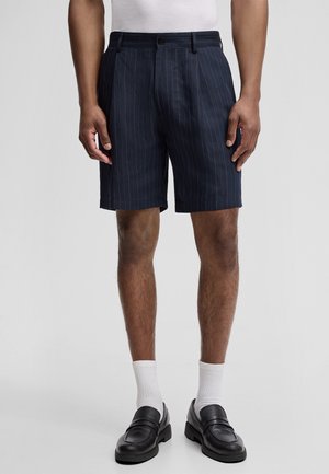 Mann trägt marineblaue Shorts mit Nadelstreifen, weiße Socken, schwarze Loafers und ein weißes Tanktop, steht vor einem einfarbigen Hintergrund.
