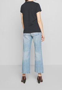 Femme se tenant de dos, portant un t-shirt noir à manches courtes, un jean droit bleu clair et des bottines à talons brunes.