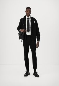 Schwarze Zip-Jacke mit Logo, weißes Hemd, schwarze Krawatte, schwarze Jogginghosen und schwarze Schnürschuhe. Minimalistisches Design, glatte Textur und taillierte Form.