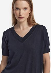 Mörkblå topp med v-ringning och kort puffärm, tillverkad av ett mjukt tyg. Har en subtil glans och elegant detaljering vid halsringningen.