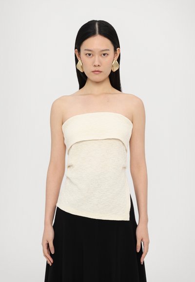 Top sans bretelles en crème avec un tissu tricoté texturé, présentant un design plié au niveau de la poitrine et un ourlet asymétrique, accompagné de grandes boucles d'oreilles en or.
