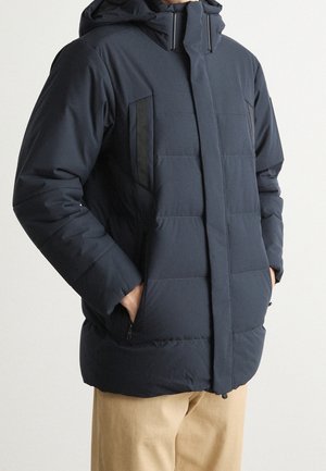 Mężczyzna ubrany w granatową, pikowaną kurtkę z kapturem i zasuwanymi na zamek bocznymi kieszeniami oraz beżowe spodnie, stojący z rękami w kieszeniach.