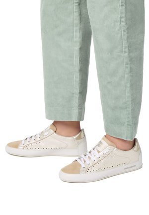 Sneaker bianchi e beige con dettagli perforati, finitura testurizzata e lacci piatti. Indossati con pantaloni in corduroy verde chiaro.