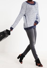 Stickad blå och vit tröja med en mjuk marinblå kant, kombinerad med grå jeans och svarta stövletter med klack, håller en svart väska.