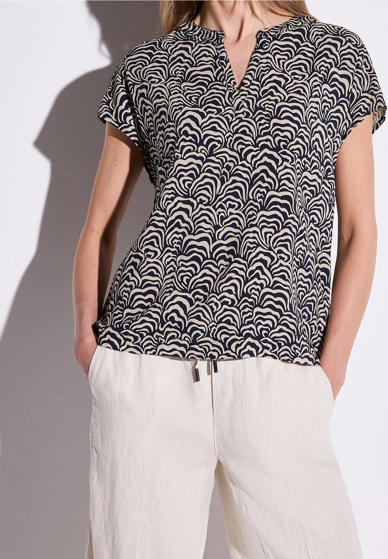 Femme portant un haut à manches courtes noir et beige à motifs avec un pantalon blanc à cordon, mains dans les poches, devant un fond uni.