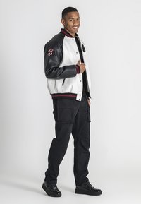 Læder- og stof varsityjakke i hvid og sort, med en rød og sort stribet krave og et ternet mærke. Sammen med sorte cargo-bukser og sneakers.