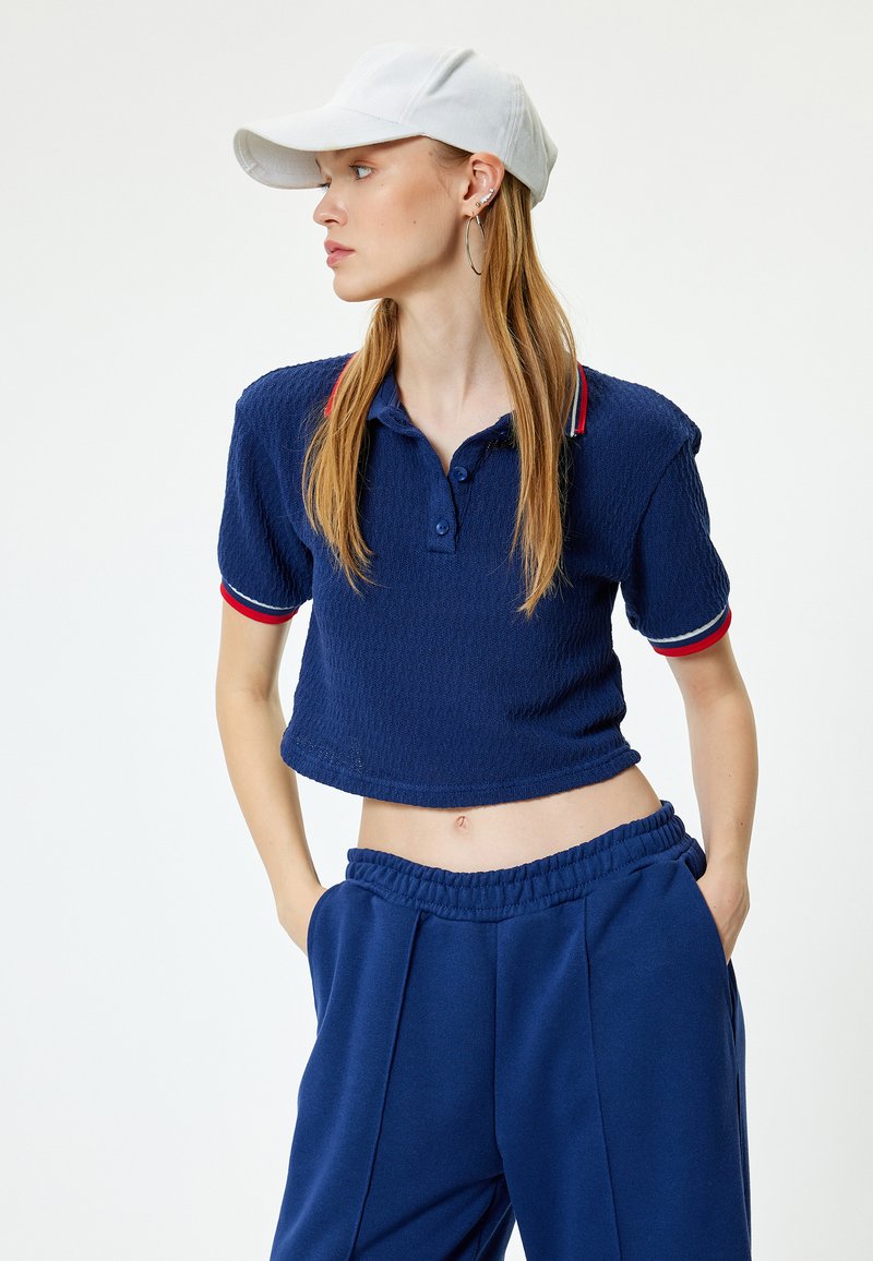 Polo shirt corto blu navy in tessuto testurizzato, colletto a contrasto rosso e bianco, abbinato a pantaloni da ginnastica blu coordinati. Cappellino bianco indossato.